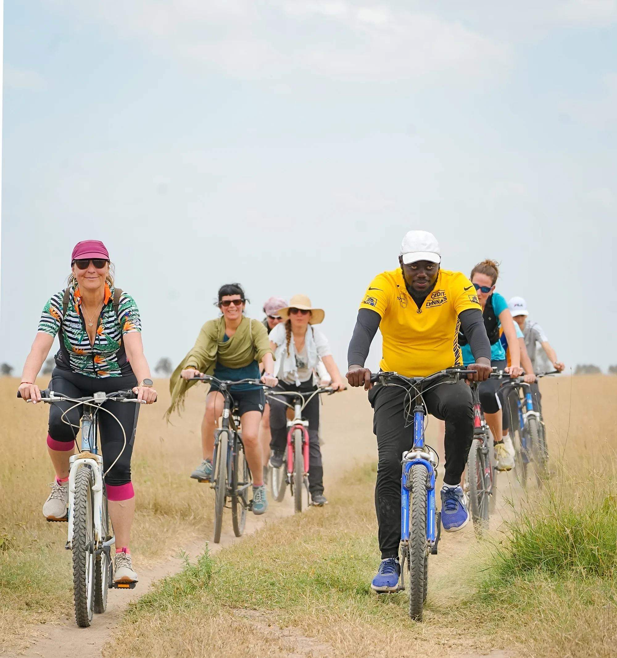 A line of cyclists rides toward the camera along a narrow dirt track through dry savannah grass, a guide in yellow among them