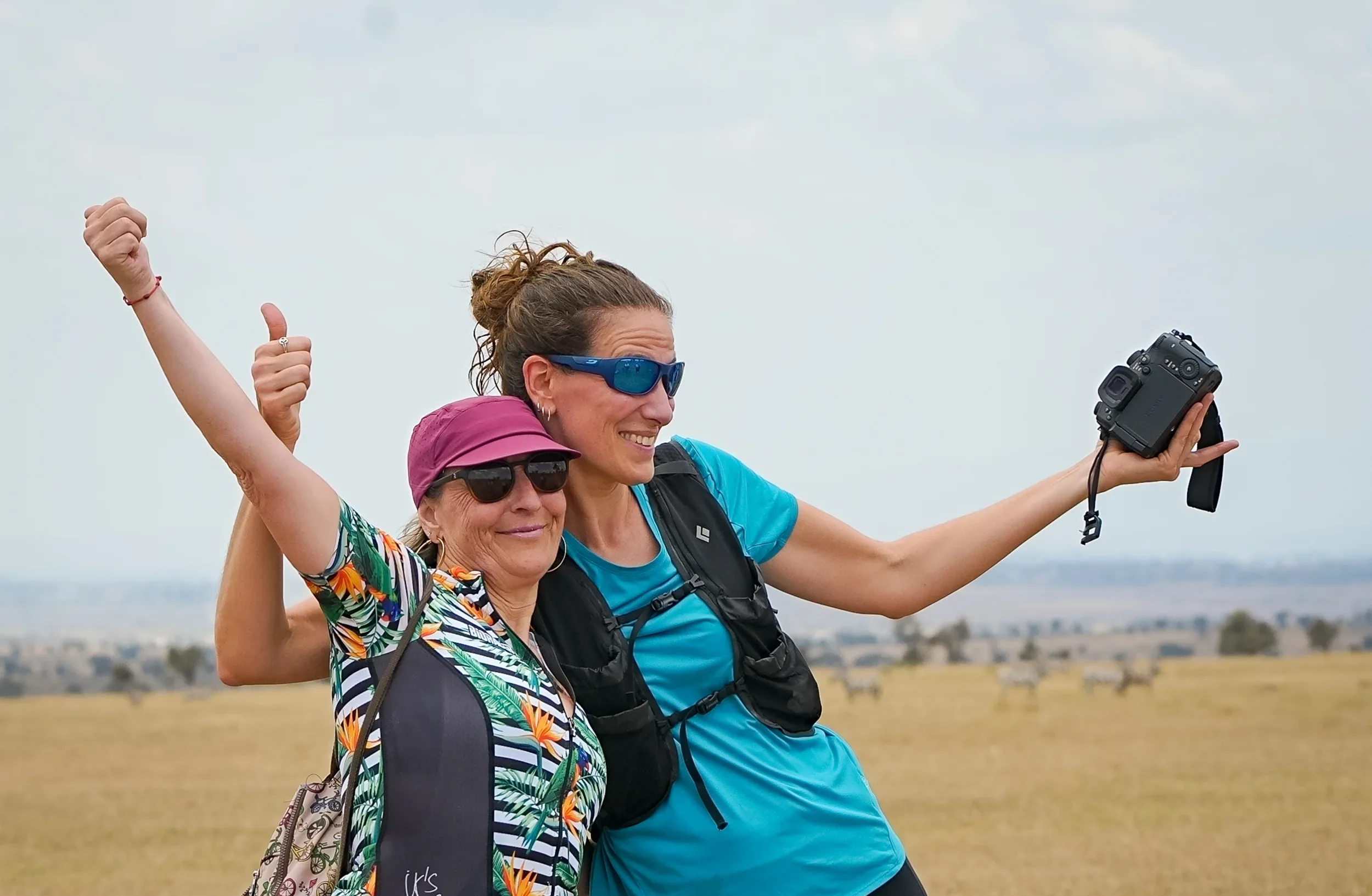Two women in cycling kit throw their arms up and cheer beside their bikes on the open plains