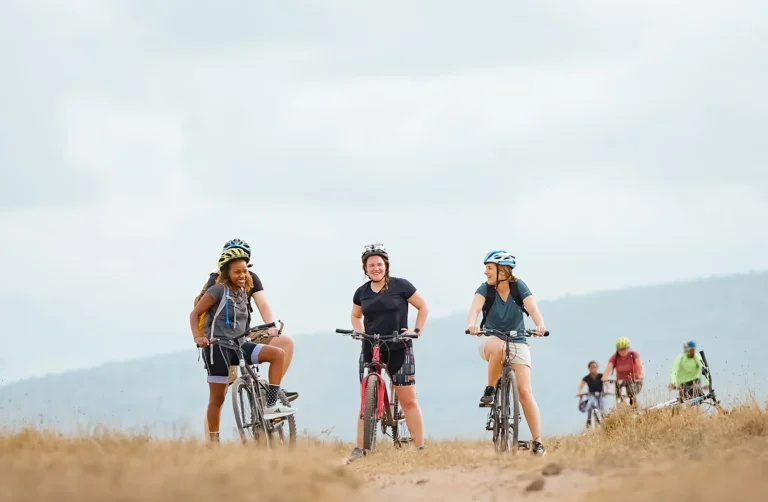 Three cyclists in helmets pause smiling on the crest of a golden grass ridge, more riders climbing behind them