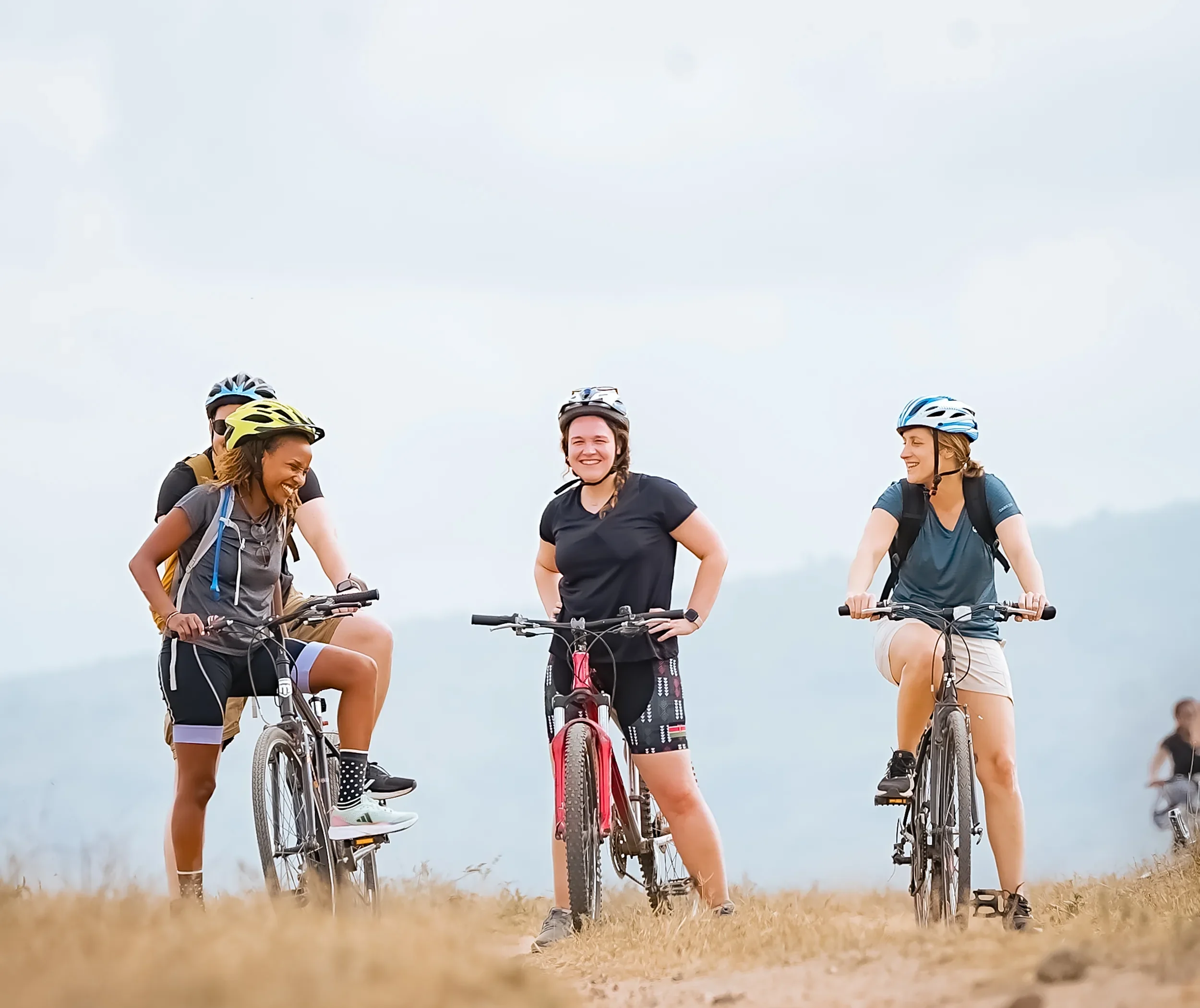 Three riders straddle their bikes on a grassy ridge, laughing together with blue hills fading behind them