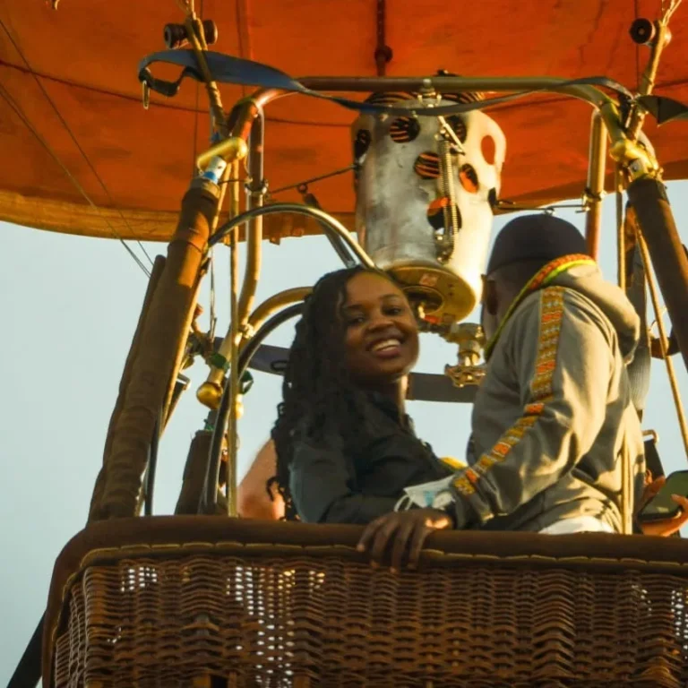 A smiling guest looks back from a wicker balloon basket as the pilot works the burner above her