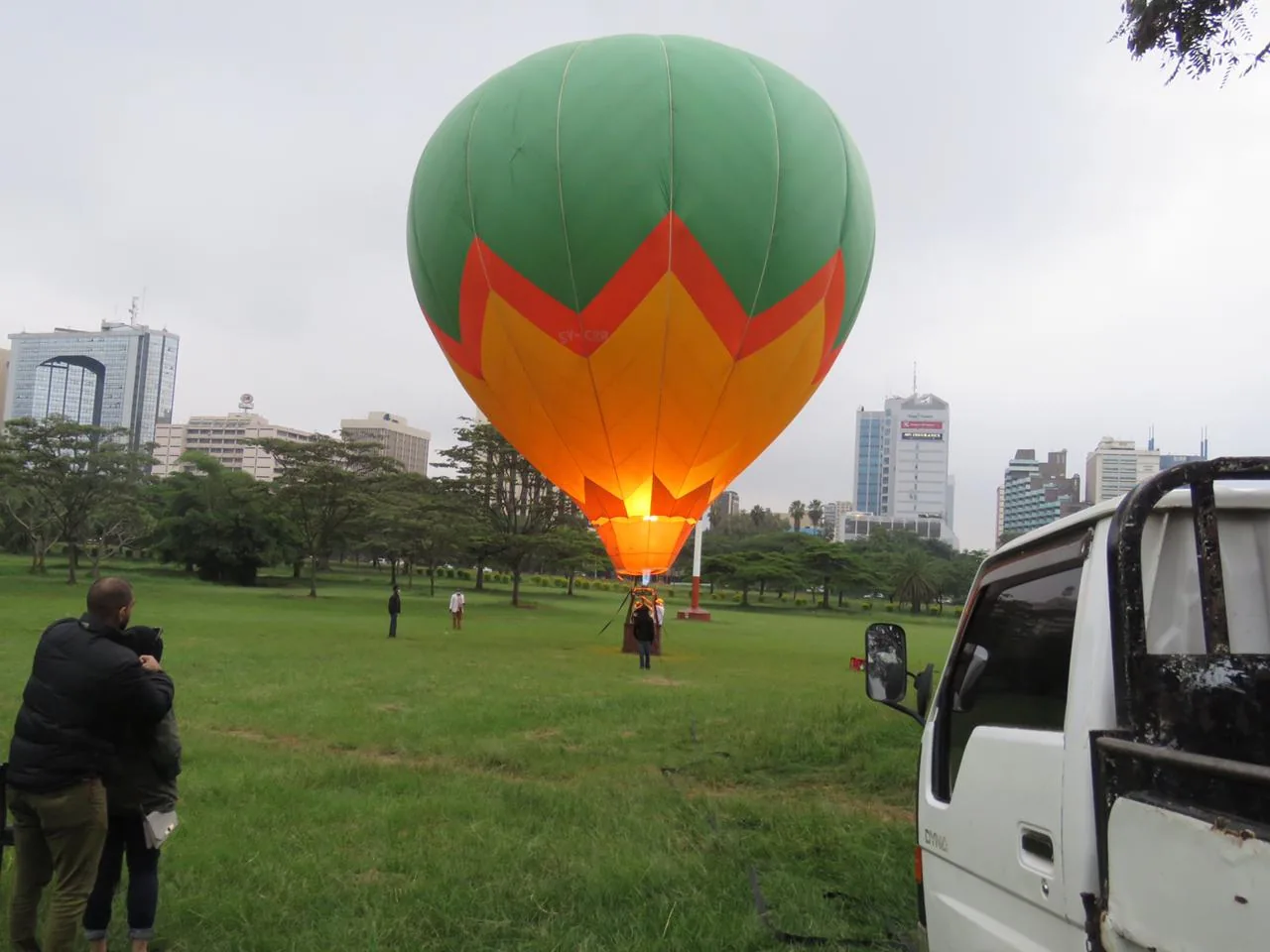 A fully inflated green and orange balloon stands on the grass of a city park, office towers rising through the trees behind