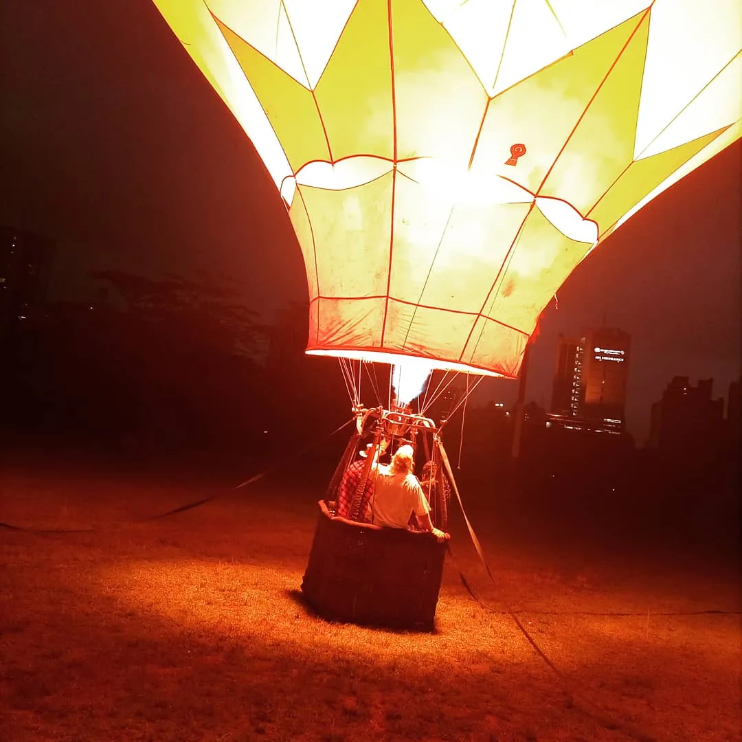 A hot air balloon glows yellow and orange from within against a dark night sky, city towers faint behind it
