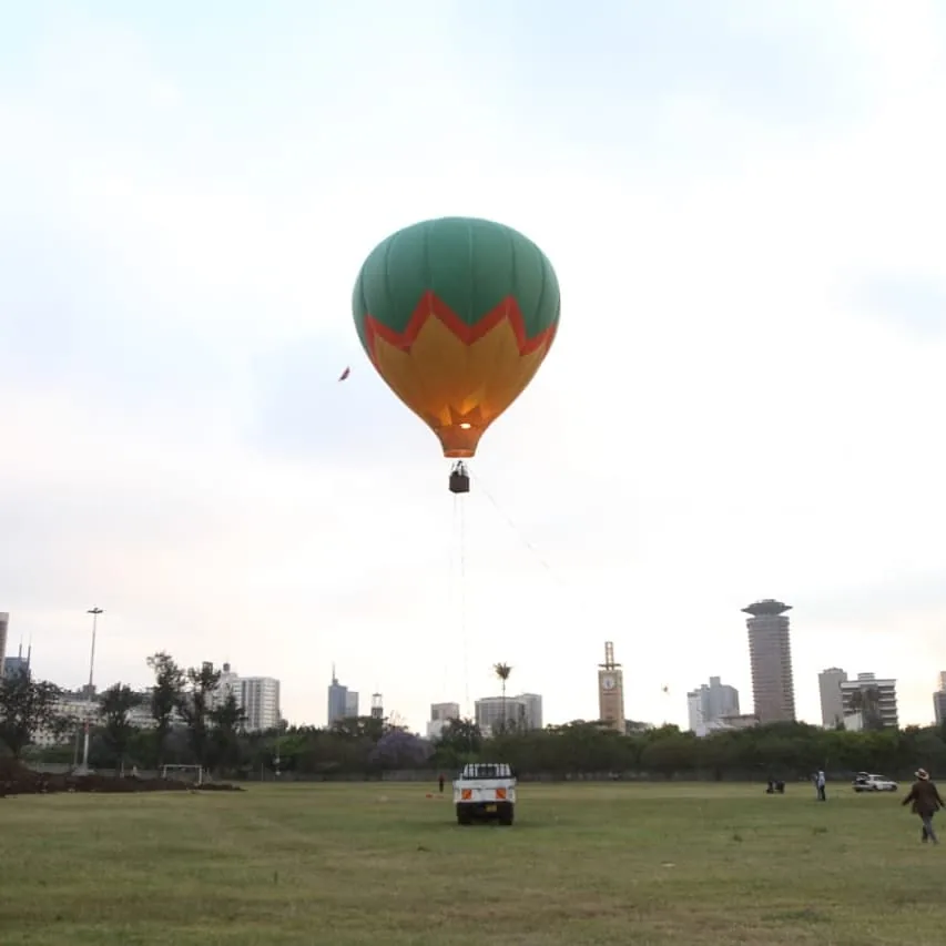 A green and orange hot air balloon hovers on its tether above the open lawns of Uhuru Park with the Nairobi skyline behind