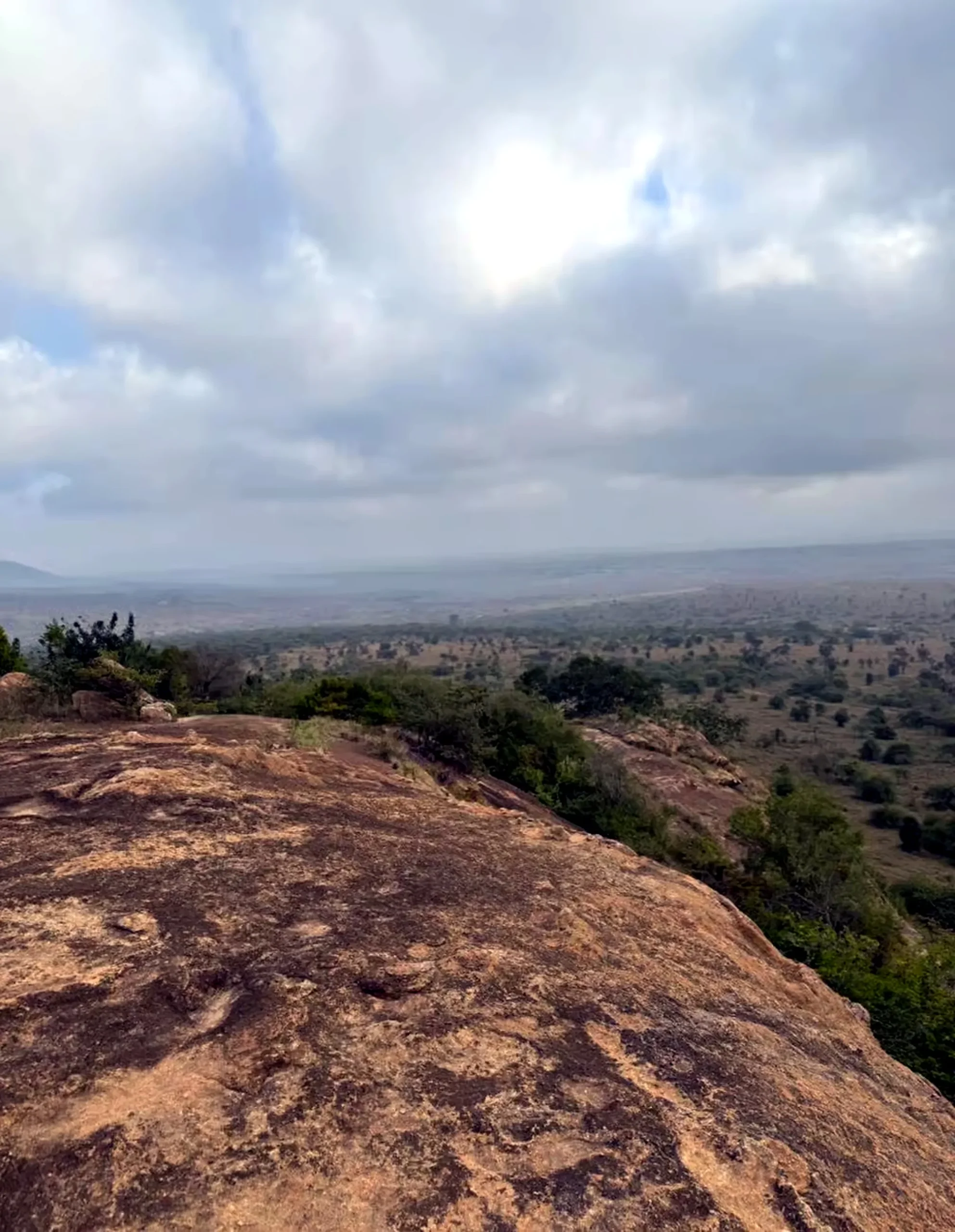 The smooth granite dome of a rocky outcrop overlooking a vast acacia-dotted plain under a bright cloudy sky