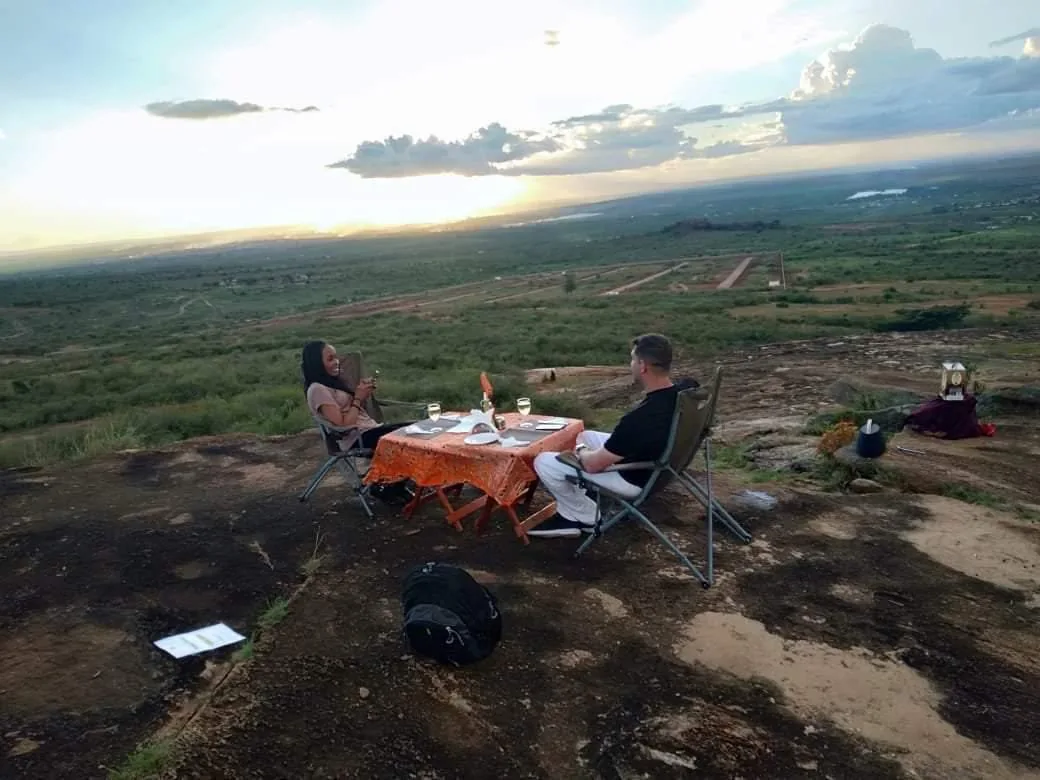 A couple sit at a small laid table on a bare rock at dusk, looking out over an immense plain streaked with sunset cloud