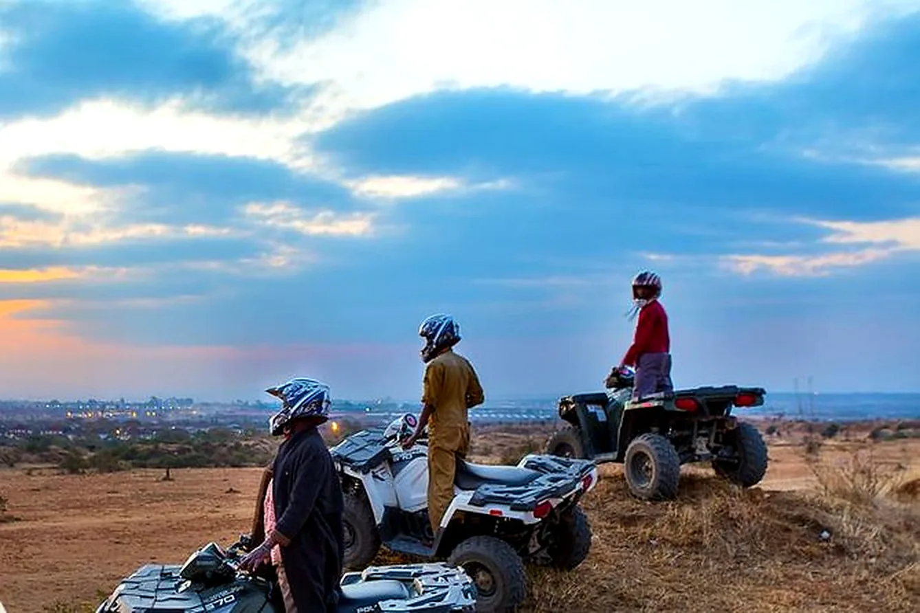 Three quad bike riders in helmets pause on a ridge at dusk, city lights glowing on the plains below near Nairobi