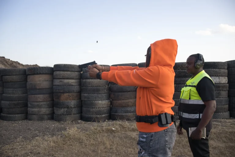 A guest fires a pistol from behind a stacked tyre barricade while an instructor watches beside her
