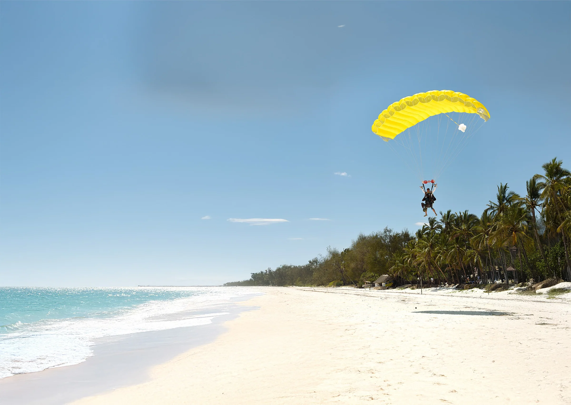 A small yellow canopy drifts down toward an empty white sand beach and turquoise sea