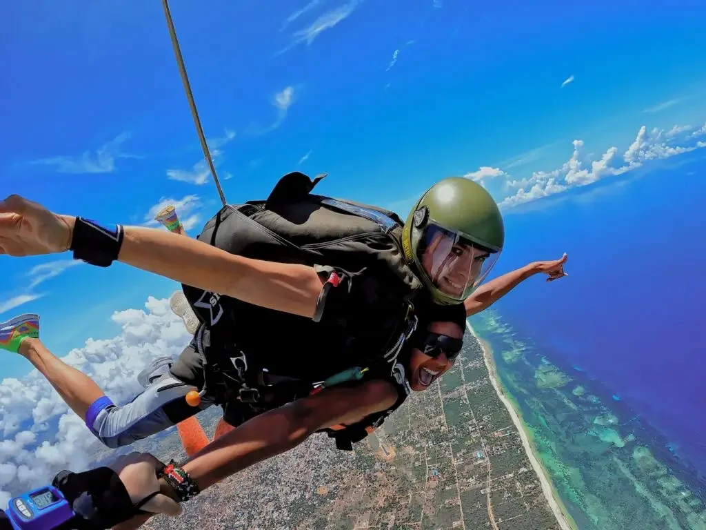 A tandem skydiving pair in freefall, arms out, with turquoise shallows and beach beneath them